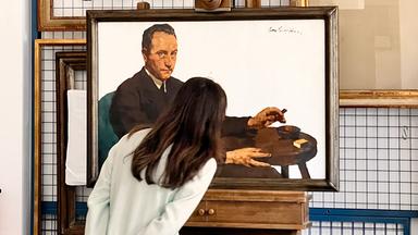 Eine Frau mit langen, dunkelbraunen Haaren steht mit dem Rücken zur Kamera und betrachtet ein Gemälde. Sie trägt einen hellen Hosenanzug und hat die Hände locker hinter ihrem Rücken verschränkt. Das Gemälde zeigt einen Mann in dunkler Kleidung, der an einem kleinen runden Tisch sitzt und eine Zigarre in der Hand hält. Neben ihm auf dem Tisch liegen weitere Gegenstände, darunter ein Streichholzbriefchen. Der Rahmen des Bildes ist aus dunklem Holz. Im Hintergrund sind weitere leere Bilderrahmen sowie ein Metallgitter zu sehen. 