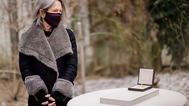 Eine Frau im Wintermantel mit Mund-Nasen-Bedeckung steht neben einem Podest, auf dem eine geöffnete Schatulle mit einer Medaille liegt.