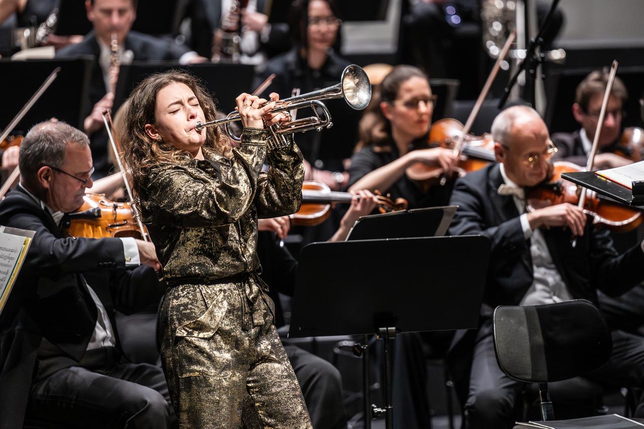 Eine junge Frau mit langen Locken, gekleidet mit einem golden glitzernden Overall, spielt vor einem großen Orchester Trompete.