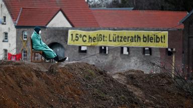 Protest gegen den Braunkohleabbau in Lützerath: Ein Demonstrant sitzt auf einem Stuhl, der auf einem Erdhaufen steht. Dahinter hängt ein Banner an einer Häuserwand, auf dem steht: "1,5 Grad Celsius heißt: Lützerath bleibt!"