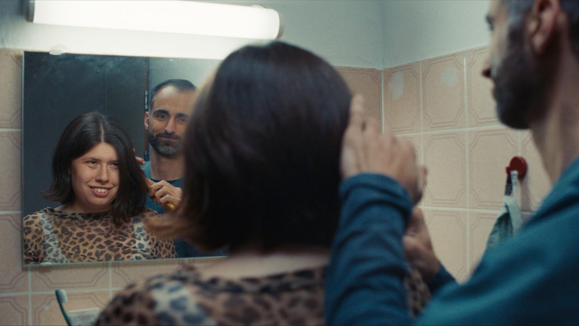 Ein Mann kämmt einer jungen Frau die Haare vor einem Badspiegel. Man sieht die Personen von hinten und  ihr Spiegelbild´.