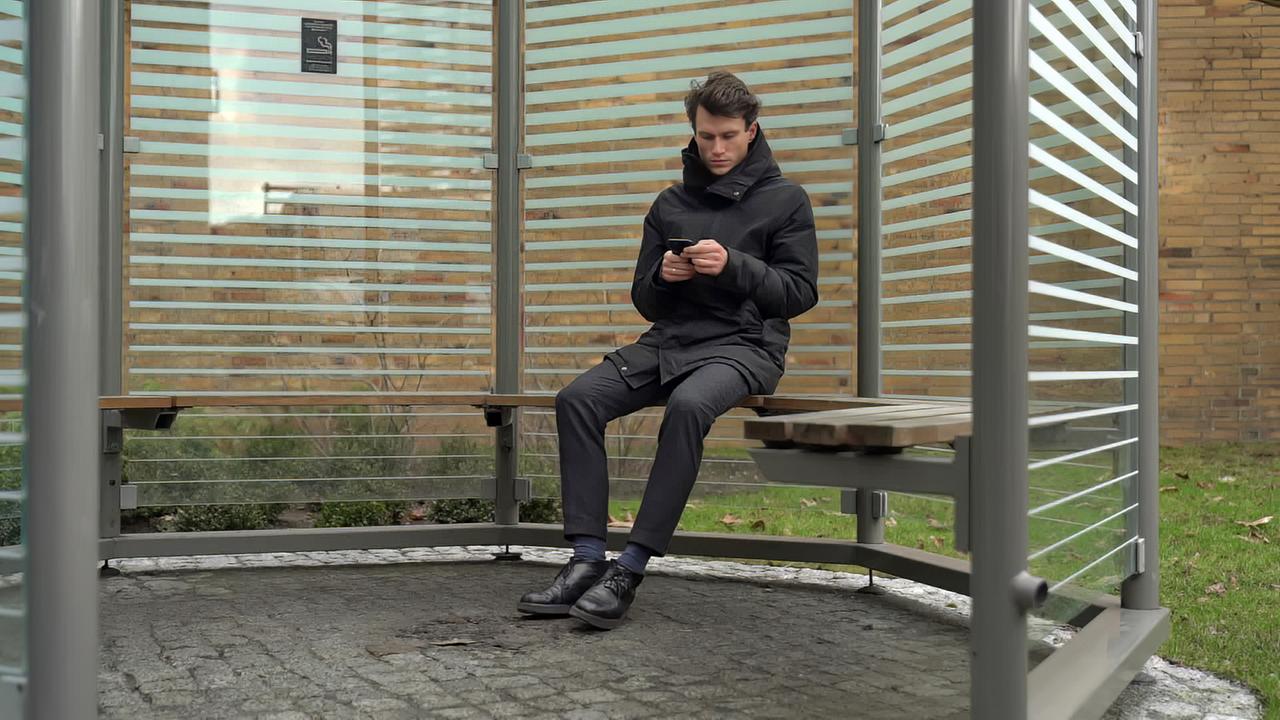 Mann sitzt auf einem Sitz in einer Haltestelle und schaut auf sein Mobiltelefon