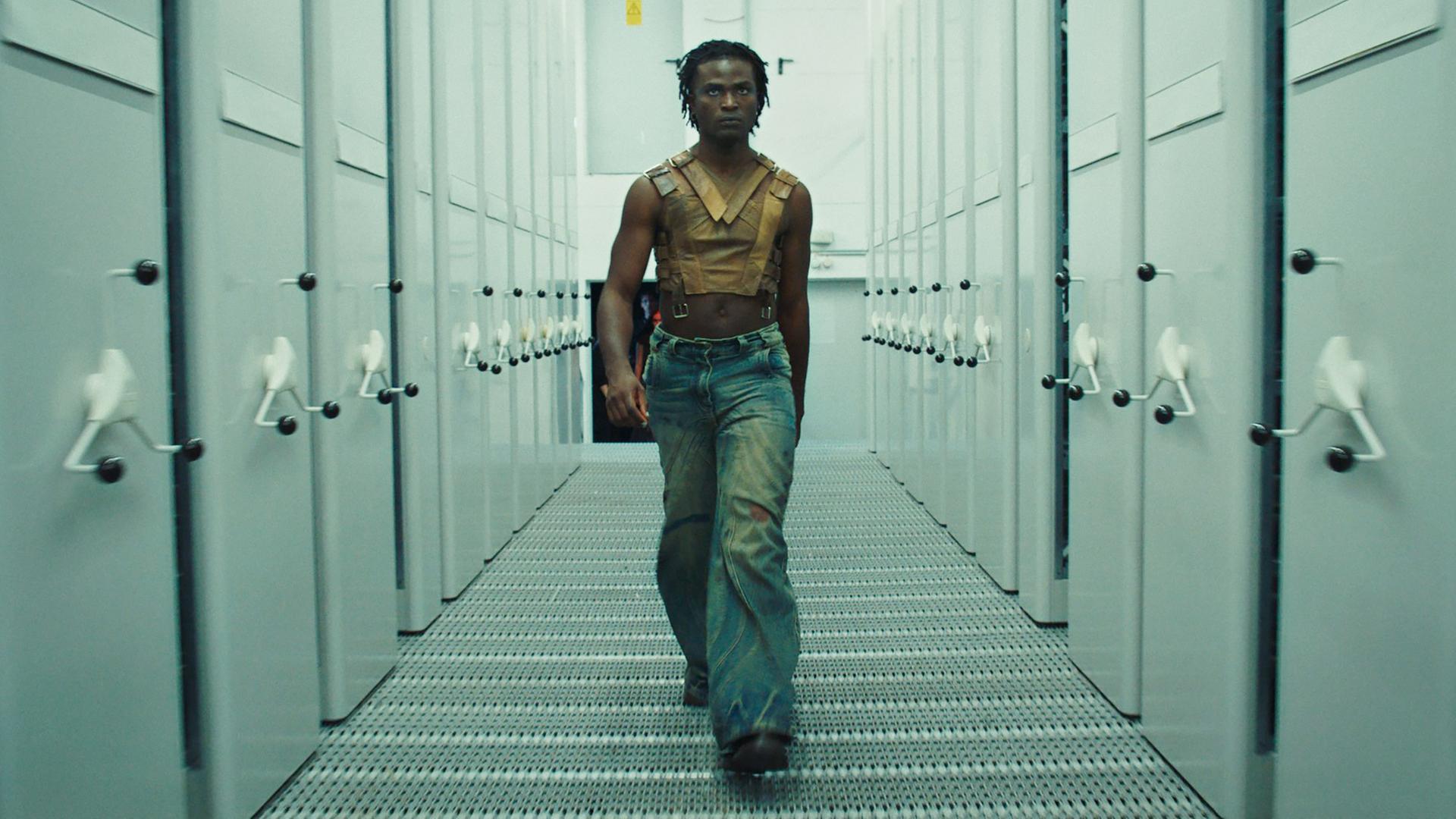  Das Bild zeigt eine Person, die selbstbewusst einen Korridor entlanggeht. Der Korridor ist von großen, grauen Schränken gesäumt, die auf beiden Seiten stehen. Die Person trägt ein bauchfreies, ärmelloses Oberteil aus braunem Leder mit Riemen und eine weit geschnittene Jeans. Das Licht ist gedämpft und erzeugt eine kühle Atmosphäre, die durch die grauen Wände und den metallenen Boden verstärkt wird. Die Person schaut direkt nach vorne, während sie auf die Kamera zugeht. 