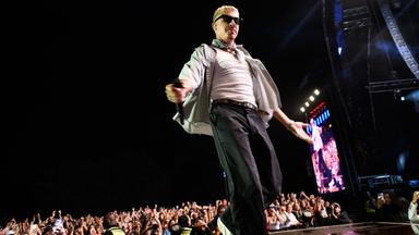 Mann mit Sonnenbrille steht abends auf einer Bühne vor großem Publikum und performt.