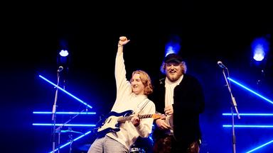 Maël & Jonas, zwei junge Männer, stehen auf einer Bühne, die im Hintergrund zum Teil blau beleuchtet ist. Jonas ist bekleidet mit hellem Pullover, hat eine E-Gitarre in der Hand und reckt die andere Hand nach oben. Maël, mit Bart und lockigen Haaren, steht neben ihm. Er trägt eine schwarze Mütze, eine schwarze Jacke und darunter ein helles Oberteil und lacht in die Kamera.