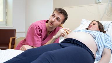 Casper Jaginiak, ein junger Mann in Krankenhauskleidung, hört mit einem Hörrohr (Stethoskop) den Bauch einer Frau ab, die auf einer Liege vor ihm liegt.