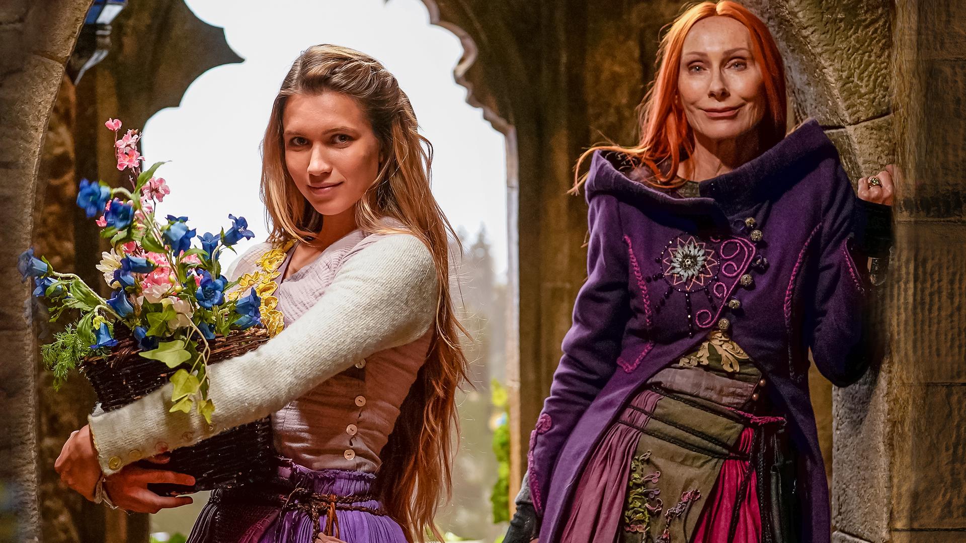 In einem alten, steinernen Gemäuer stehen Rapunzel, gespielt von Anne-Lena Schwing, und Zauberin Eleonor, gespielt von Andrea Sawatzi, vor einem steinernen Fensterrahmen und schauen in die Kamera.