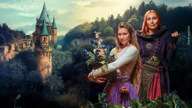 In einem alten, steinernen Gemäuer stehen Rapunzel, gespielt von Anne-Lena Schwing, und Zauberin Eleonor, gespielt von Andrea Sawatzi, vor einem steinernen Fensterrahmen und schauen in die Kamera.