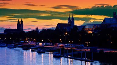 Dunkle Umrisse einer Stadt an einem Fluss vor orange-rot gefärbertem Himmel, Lichter spiegeln sich im Wasser.