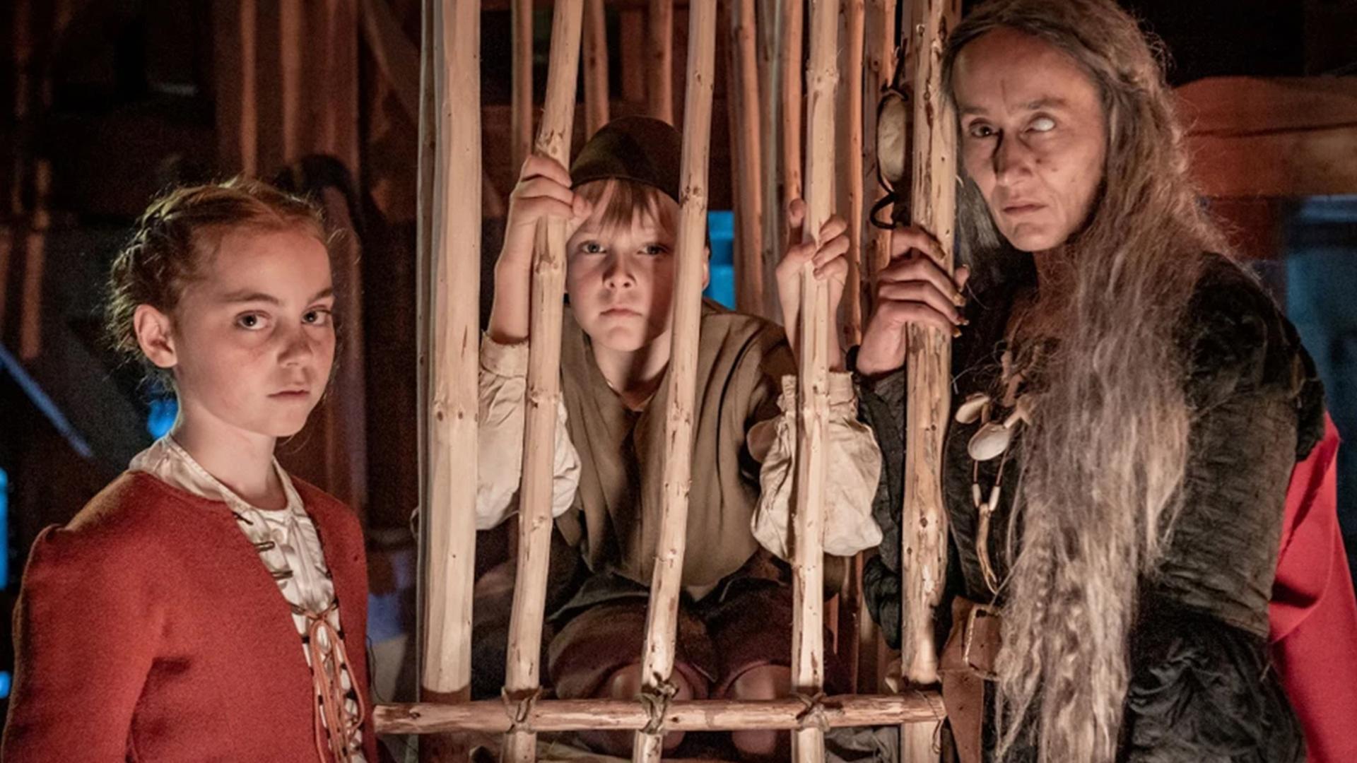 Spielszene: Hänsel sitzt im Holzkäfig, rechts von ihm steht Gretel, links von ihm eine Hexe