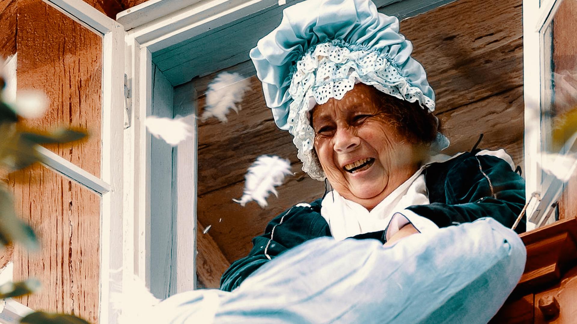 Frau Holle schüttelt ein Kissen aus dem Fenster heraus. 