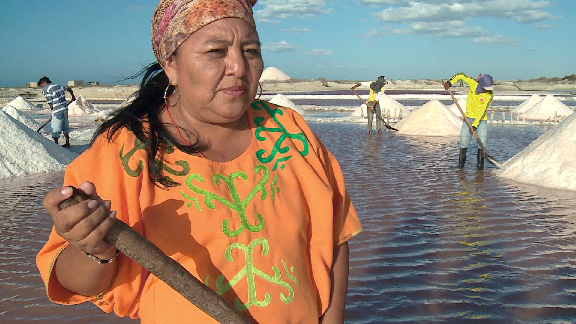Luz Marina steht mit eine Schaufel in einer Saline. Hinter ihr Mitarbeiter und Salzhügel.