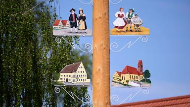 Detail eines Maibaums. Rechts und links vom Stamm sind bemalte Schilder mit Personen in historischen Kleidern und Gebäude aus dem Ort zu sehen.