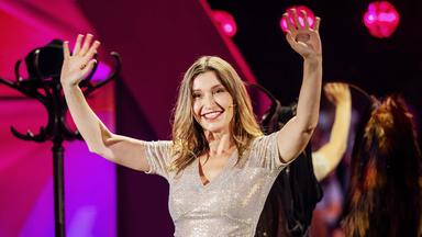 Malarina trägt ein Shirt sowie eine Hose die glitzert. Sie steht in der Mitte des Bildes und hält beide Hände nach oben. Im Hintergrund ist ein Jackenständer zu sehen. 