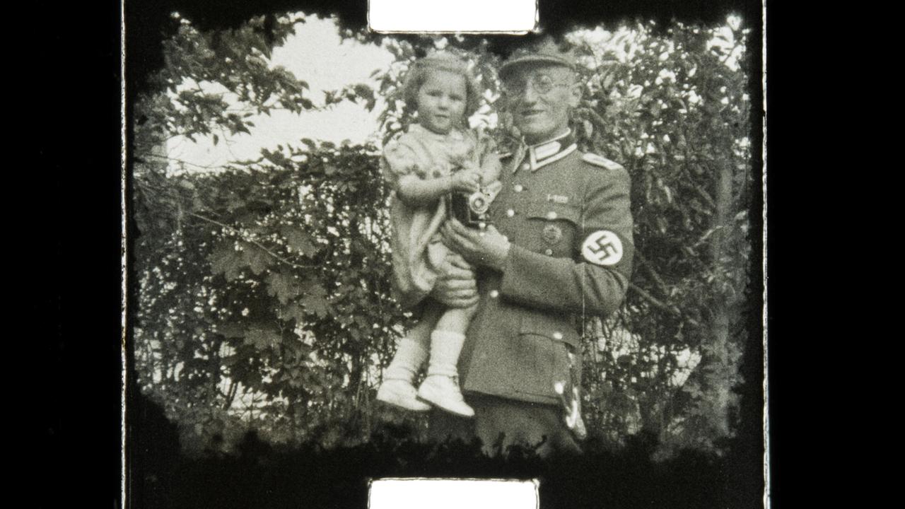 Ein Mann in Nazi-Uniform hält ein kleines Mädchen im Sommerkleid auf dem Arm