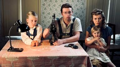 Ein Mann und eine Frau sitzen mit ihren Kindern an einem Tisch und schauen einen Film. Kleine Filmspulen und das Filmvorführgerät stehen auf dem Tisch vor ihnen.