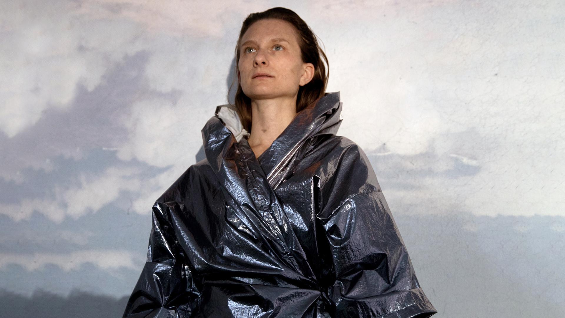 Eine Frau mit blau glänzender Jacke vor einem grauen Hintergrund.