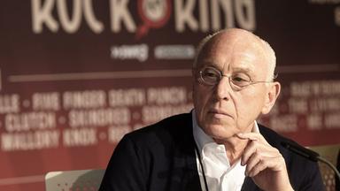 Ein älterer Mann mit grauen Haaren und Metallbrille vor einem "Rock am Ring"-Plakat