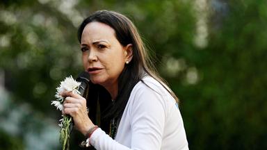 Eine Frau mit langen Haaren spricht in ein Mikrofon, das sie zusammen mit einer weißen Blume in der Hand hält.