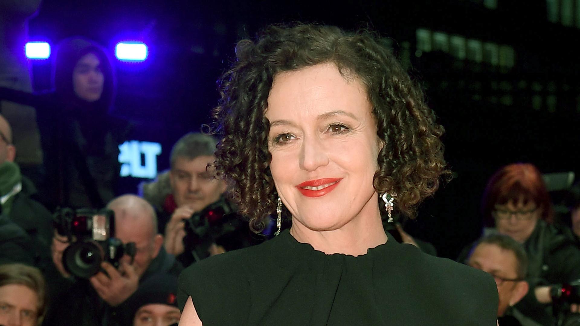 Eine Frau mit dunklen lockigen Haaren und rotem Lippenstift lächelt in die Kamera.