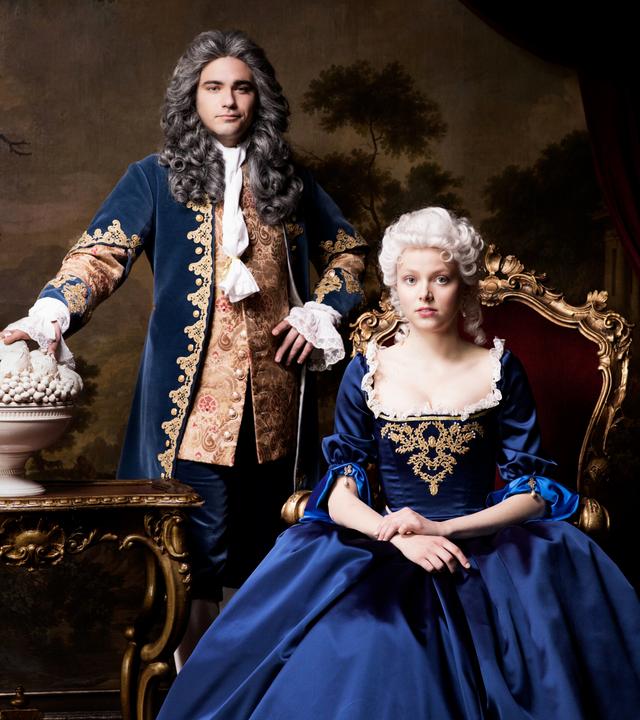 Ein Mann steht neben einer sitzenden Frau in blauer Robe.