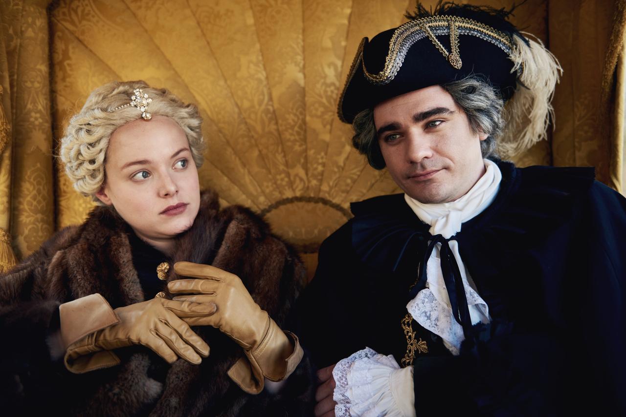 Das Bild zeigt zwei historische Figuren in einem eleganten, opulenten Setting. Links im Bild sitzt eine Frau mit blonden, lockigen Haaren, die kunstvoll frisiert sind. Sie trägt ein braunes, pelzverziertes Cape und beige Handschuhe. Ihr Gesichtsausdruck ist nachdenklich, und sie blickt leicht zur Seite.  Rechts neben ihr sitzt ein Mann, der eine schwarze, reich verzierte Kleidung trägt. Er hat ebenfalls graue Haare und trägt einen auffälligen Hut mit Verzierungen. Sein Gesichtsausdruck wirkt gelassen, während er die Frau ansieht.  Im Hintergrund ist ein goldener Stoff zu sehen, der die opulente Atmosphäre des Moments unterstreicht. Die gesamte Szene vermittelt ein Gefühl von historischem Flair und Intensität in der Darstellung der Charaktere.