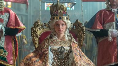 Das Bild zeigt eine Szene aus der Geschichte von Maria Theresia. Im Mittelpunkt sitzt eine junge Frau, die Maria Theresia darstellt, auf einem prunkvollen, goldenen Thron. Sie trägt ein aufwendiges Kostüm mit viel Spitze und Edelsteinen sowie einen prächtigen, auffälligen Hut oder eine Krone auf dem Kopf. Ihr Gesichtsausdruck ist ernst und konzentriert.  Sie ist umgeben von mehreren Personen, die in historischen Gewändern gekleidet sind. Diese Personen stehen zu beiden Seiten des Thrones und scheinen offizielle Würdenträger oder Berater zu sein. Im Hintergrund sind Ansichten von farbigen Fenstern erkennbar, die dem Raum eine feierliche Atmosphäre verleihen.  Die gesamte Szenerie wirkt majestätisch und historisch, was durch die aufwändigen Details der Kleidung und die Gestaltung des Thrones verstärkt wird.