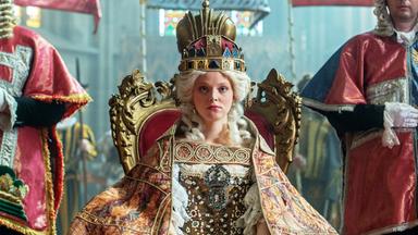 Das Bild zeigt eine Szene aus der Geschichte von Maria Theresia. Im Mittelpunkt sitzt eine junge Frau, die Maria Theresia darstellt, auf einem prunkvollen, goldenen Thron. Sie trägt ein aufwendiges Kostüm mit viel Spitze und Edelsteinen sowie einen prächtigen, auffälligen Hut oder eine Krone auf dem Kopf. Ihr Gesichtsausdruck ist ernst und konzentriert.  Sie ist umgeben von mehreren Personen, die in historischen Gewändern gekleidet sind. Diese Personen stehen zu beiden Seiten des Thrones und scheinen offizielle Würdenträger oder Berater zu sein. Im Hintergrund sind Ansichten von farbigen Fenstern erkennbar, die dem Raum eine feierliche Atmosphäre verleihen.  Die gesamte Szenerie wirkt majestätisch und historisch, was durch die aufwändigen Details der Kleidung und die Gestaltung des Thrones verstärkt wird.