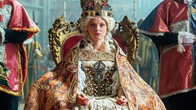 Das Bild zeigt eine Szene aus der Geschichte von Maria Theresia. Im Mittelpunkt sitzt eine junge Frau, die Maria Theresia darstellt, auf einem prunkvollen, goldenen Thron. Sie trägt ein aufwendiges Kostüm mit viel Spitze und Edelsteinen sowie einen prächtigen, auffälligen Hut oder eine Krone auf dem Kopf. Ihr Gesichtsausdruck ist ernst und konzentriert.  Sie ist umgeben von mehreren Personen, die in historischen Gewändern gekleidet sind. Diese Personen stehen zu beiden Seiten des Thrones und scheinen offizielle Würdenträger oder Berater zu sein. Im Hintergrund sind Ansichten von farbigen Fenstern erkennbar, die dem Raum eine feierliche Atmosphäre verleihen.  Die gesamte Szenerie wirkt majestätisch und historisch, was durch die aufwändigen Details der Kleidung und die Gestaltung des Thrones verstärkt wird.