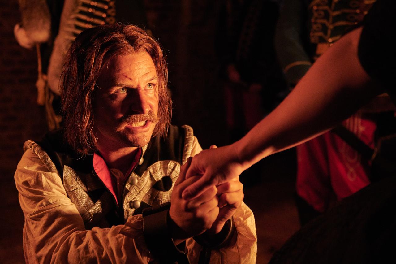 Das Bild zeigt eine Szene aus dem historischen Drama "Maria Theresia". Im Vordergrund ist ein Mann mit langen, lockigen, braunen Haaren und einem Schnurrbart zu sehen, der in einem prächtigen historischen Kostüm gekleidet ist. Er hat ein ernsthaftes und eindringliches Gesicht und schaut direkt zur Kamera oder zu einer anderen Person, während er eine Hand hält. Sein Blick vermittelt eine Mischung aus Entschlossenheit und Verzweiflung.  Die Beleuchtung ist gedämpft und wirkt warm, was die dramatische Stimmung der Szene verstärkt. Im Hintergrund sind schemenhaft weitere Figuren zu erkennen, die ebenfalls in historischem Militärgewand gekleidet sind. Der Hintergrund ist unscharf, was den Fokus auf die handelnde Person im Vordergrund verstärkt.  Insgesamt vermittelt das Bild eine spannungsgeladene Atmosphäre, die auf eine bedeutende und emotionale Interaktion im Kontext der Handlung hinweist.