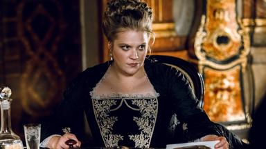 Das Bild zeigt eine Frau in einem historischen Raum, die an einem Tisch sitzt. Sie trägt ein elegantes schwarzes Kleid mit aufwendigen, hellen Stickereien im oberen Bereich. Ihr Haar ist hochgesteckt, und sie hat große, auffällige Ohrringe. Die Mimik der Frau wirkt ernst und nachdenklich, sie scheint auf etwas konzentriert zu sein, während sie auf einem Blatt Papier schaut, das auf dem Tisch vor ihr liegt.   Auf dem Tisch stehen ein Glas Wasser und eine Karaffe, sowie einige Dokumente. Der Hintergrund zeigt Wände mit reich verzierten Holzvertäfelungen und Möbel, die auf eine historische Epoche hinweisen. Die Atmosphäre wirkt majestätisch und zeugt von einem prunkvollen Interieur.