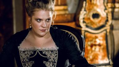 Das Bild zeigt eine Frau in einem historischen Raum, die an einem Tisch sitzt. Sie trägt ein elegantes schwarzes Kleid mit aufwendigen, hellen Stickereien im oberen Bereich. Ihr Haar ist hochgesteckt, und sie hat große, auffällige Ohrringe. Die Mimik der Frau wirkt ernst und nachdenklich, sie scheint auf etwas konzentriert zu sein, während sie auf einem Blatt Papier schaut, das auf dem Tisch vor ihr liegt.   Auf dem Tisch stehen ein Glas Wasser und eine Karaffe, sowie einige Dokumente. Der Hintergrund zeigt Wände mit reich verzierten Holzvertäfelungen und Möbel, die auf eine historische Epoche hinweisen. Die Atmosphäre wirkt majestätisch und zeugt von einem prunkvollen Interieur.