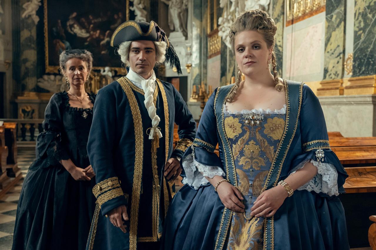 Auf dem Bild befinden sich drei Personen in einem historischen Raum, der durch hohe Decken und umfangreiche Dekorationen geprägt ist. Im Hintergrund sind Elemente wie große Gemälde und Holzvertäfelungen sichtbar.   Links steht eine Frau in einem schwarzen, eleganten Kleid mit langen Ärmeln und einem feinen Halsband. In der Mitte steht ein Mann in einer blauen, mit goldenen Verzierungen versehenen Jacke, unter der er ein weißes Hemd mit einem hohen Kragen trägt. Er hat eine aufwendige Perücke und scheint ernst und nachdenklich zu sein. Rechts ist eine Frau in einem prunkvollen, blauen Kleid zu sehen, das ebenfalls mit goldenen Mustern verziert ist. Sie trägt Schmuck und hat ihre Haare hochgesteckt.  Die Atmosphäre des Bildes vermittelt einen Eindruck von Macht und Ehrfurcht, passend zu einer königlichen Umgebung.