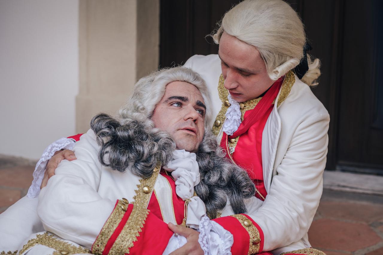 Das Bild zeigt zwei Männer in historischer Kleidung, die im Stil des 18. Jahrhunderts gestaltet sind. Der erste Mann, auf der linken Seite, trägt eine prächtige weiße Jacke mit goldenen Verzierungen und hat eine voluminöse, graue Perücke mit großen Locken. Sein Gesichtsausdruck wirkt ernst und nachdenklich.  Der zweite Mann, auf der rechten Seite, trägt ebenfalls eine elegante weiße Jacke, welche mit roten Akzenten und goldenen Knöpfen verziert ist. Sein Gesicht hat einen besorgten Ausdruck, während er den ersten Mann hält. Die beiden scheinen in einer emotionalen Interaktion zu stehen.  Im Hintergrund ist eine unauffällige Wand zu sehen, die in neutralen Farben gehalten ist. Der Boden ist mit großen, terrakottafarbenen Fliesen ausgelegt. Die Szene wirkt ruhig und ergreifend, mit einem starken Fokus auf die dargestellten Charaktere.