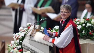 Eine Pfarrerin, gekleidet in eine rote Robe, steht auf einer Blumengeschmückten Kanzel in einer Kirche.