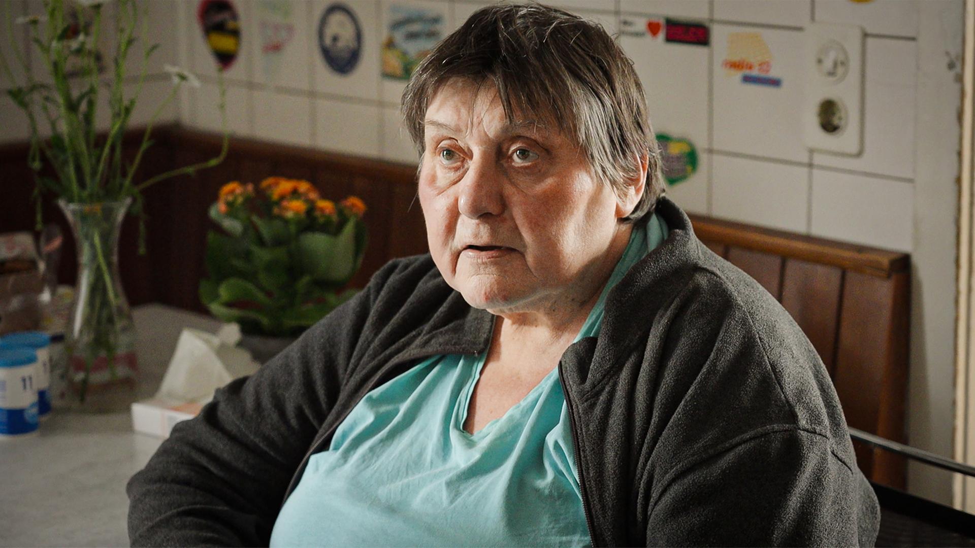 Man sieht eine ältere Frau die in der Küche sitzt und geistesabwesend an der Kamera vorbeischaut.