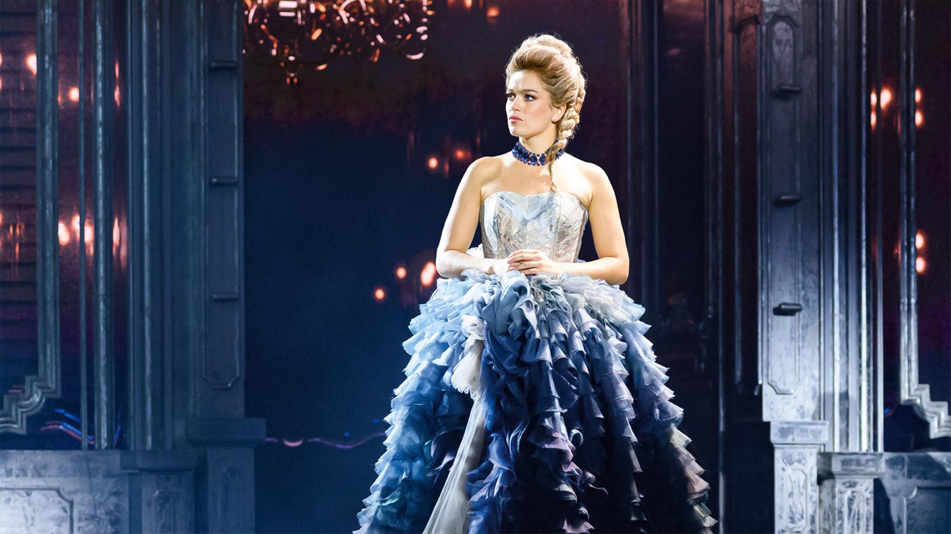 Das Bild zeigt eine Szene aus einem Musical, das sich mit dem Leben von Maria Theresia beschäftigt. Im Vordergrund steht eine weibliche Figur, die eine opulente, mehrschichtige, blau-weiße Robe trägt. Der Unterteil der Robe besteht aus vielen Rüschen, die in verschiedenen Blau- und Grautönen gestaltet sind. Die Figur hat eine geflochtene Frisur und trägt eine auffällige Halskette. Ihr Gesichtsausdruck ist ernst und nachdenklich.   Im Hintergrund sind dunkle Wände zu sehen, die mit eleganten, hochglänzenden Oberflächen und einem dezenten Lichtspiel akzentuiert sind. Einige Lichtquellen scheinen aus dem Hintergrund zu strahlen, was eine dramatische Atmosphäre schafft. Die Szene vermittelt ein Gefühl von Eleganz und Stärke, unterstreicht die historische Thematik des Musicals und reflektiert die Herausforderungen einer starken Frau in einer Machtposition.
