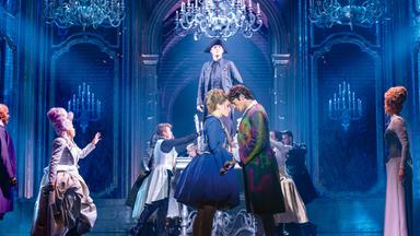 Das Bild zeigt eine Szene aus dem Musical "Maria Theresia - First Working Mom". Die Bühne ist prächtig dekoriert, mit großen, eleganten Kronleuchtern und einem opulenten Hintergrund, der an historische Architektur erinnert. In der Mitte der Szene stehen mehrere Darsteller.   Eine Frau in einem blauen Kleid steht nahe einem Mann, der farbenfrohe Kleidung trägt. Sie scheinen sich anzusehen, während hinter ihnen weitere Darsteller in anspruchsvollen Kostümen und einer dramatischen Pose in Aktion sind. Im Hintergrund steht ein Mann in schwarzer Kleidung auf einer erhöhten Plattform. Die Beleuchtung ist intensiv und schafft eine dramatische Atmosphäre, mit bläulichen Lichtern, die auf die Bühne scheinen.  Das Bild vermittelt eine dynamische und emotionale Stimmung, die das Thema des Musicals widerspiegelt, das sich mit dem Leben von Maria Theresia, einer herausragenden historischen Figur, befasst.