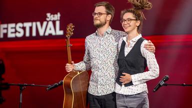 Marie Diot steht auf der rechten Seite des Bildes. Auf der linken Seite ist ihr Gitarrist zu sehen. Der Hintergrund ist rot
