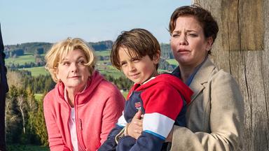 Eine Frau sitzt mit einem Jungen auf dem Schoß auf einer Bank, daneben eine ältere Frau. Zwei weitere Personen stehen daneben, alle blicken ernst. Der Hintergrund zeigt eine grüne Wiese und eine weite Landschaft.