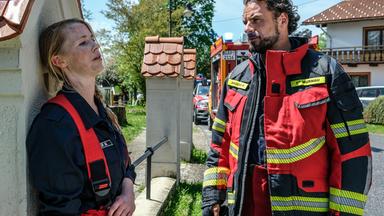 "Marie fängt Feuer - Die Feuertaufe": Marie (Christine Eixenberger) lehnt sich an eine steinerne Zauneinfriedung. Ihr gegenüber steht ihr Feuerwehrkollege Peter (Stephan Luca), der zu ihr herüberschaut.