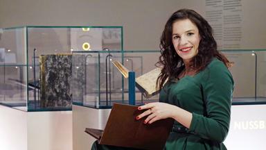 Eine dunkelhaarige Frau im grünen Kleid mit einem aufgeschlagenen Lederbuch vor Glasvitrinen.