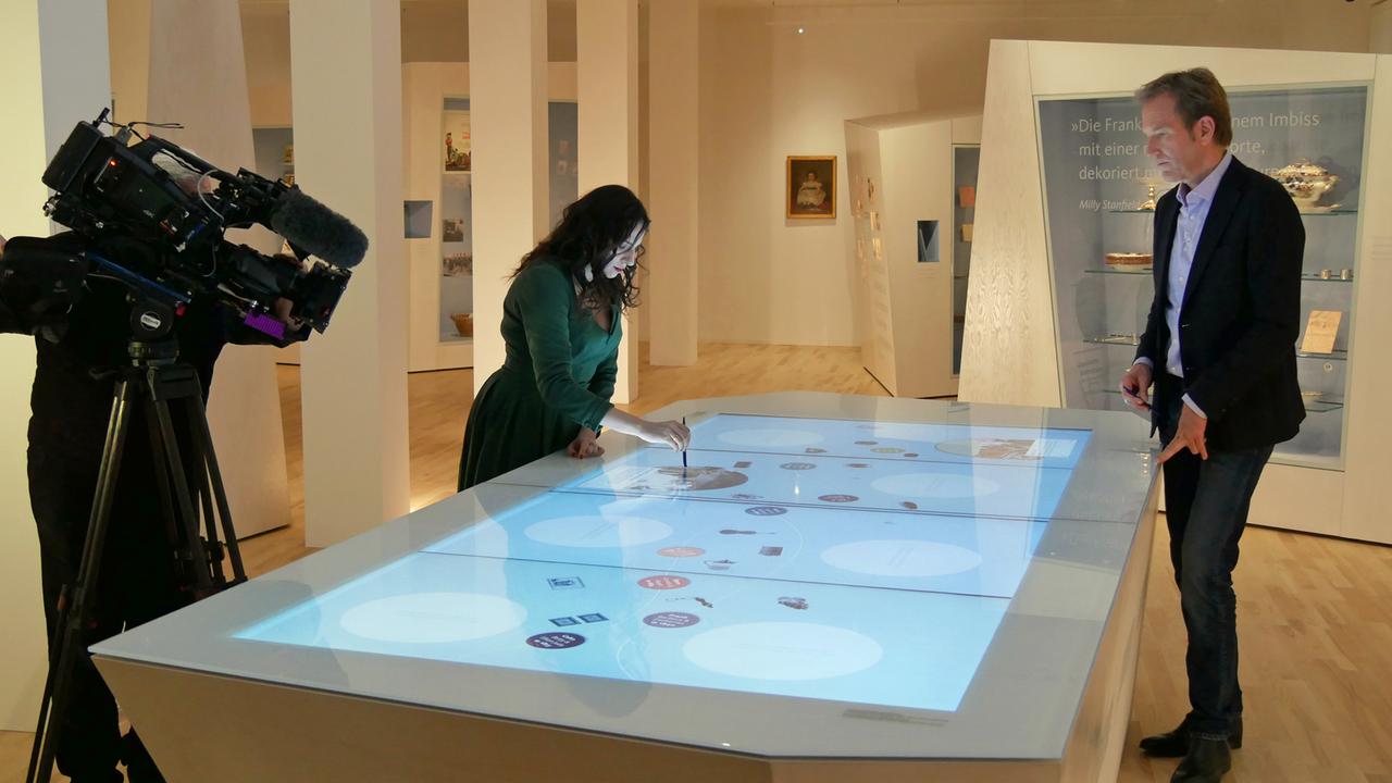 Eine Frau und der Moderator stehen neben einem großen Tisch, dessen Platte leuchtet. Die Frau berührt ein Objekt unter der Platte mit einem Stift. Daneben eine Fernsehkamera.
