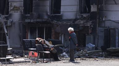 Zwei Menschen sitzen und stehen vor einem ausgebombten Haus in der Stadt Mariupol. Auf der Straße stehen Sofas und Stühle zwischen Trümmern.