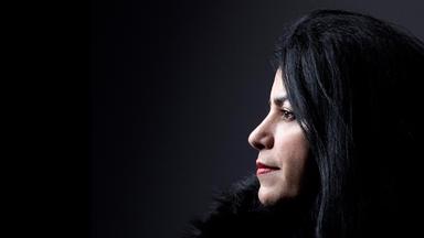 Foto von einer Frau mit langen dunklen Haaren im Profil. Die Frau trägt eine schwarze Pelzjacke und steht vor einem dunklen Hintergrund