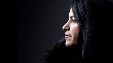 Foto von einer Frau mit langen dunklen Haaren im Profil. Die Frau trägt eine schwarze Pelzjacke und steht vor einem dunklen Hintergrund