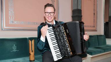 Ein junger Mann sitzt mit einem Akkordeon auf einer Bank. Neben ihm eine Trophäe aus Holz.