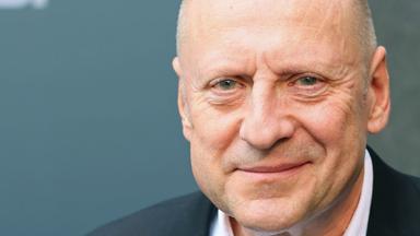 Das Gesicht eines glatzköpfigen Mannes mittleren Alters im Anzug, der dezent in die Kamera lächelt.