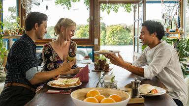 "Matze, Kebab und Sauerkraut": Noah (Franz Dinda), Charlottes (Christine Eixenberger) und Hakim (Omar El-Saeidi) sitzen gemeinsam am Tisch. Hakim hält Charlotte einen Teller hin, im Hintergrund glitzert der See