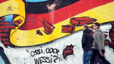 Drei verschwommene Personen gehen vor einer Mauer in Berlin vorbei, auf die eine Deutschlandfahne und einstürzendes Mauerwerk sowie der Schriftzug "Ossi oder Wessi?" gesprayt sind.